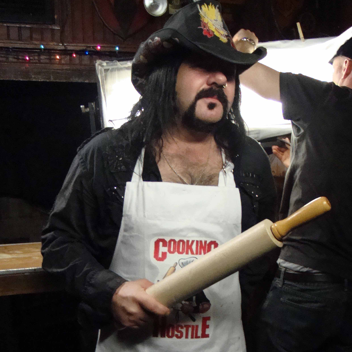 Person wearing a black hat and apron with 'Cooking is Hustle' text, holding a rolling pin.